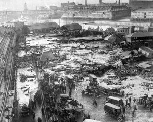 An aerial shot of a neighborhood with most buildings reduced to rubble. The harbor is visible in the distance. A crowd of people and ambulances loiter at the edge of a shallow lake of liquid, with more  walking on it and searching the wreckage.