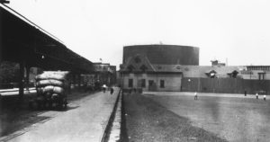 A black-and-white photo of a neighborhood. A bare playground is on the right and an elevated train track is on the left. In the distance, a huge cylindrical tank looms over a row of buildings.